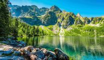 Widok na Morskie Oko, jezioro otoczone górami w Tatrach. Woda ma niebiesko-zielony kolor. Dookoła jeziora znajdują się sosny, skały oraz góry.
