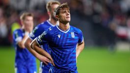  Robert Gumny w niebieskiej koszulce Lecha Poznań z rękami na biodrach, stojący na boisku piłkarskim z zamyślonym, spoglądającym w górę wyrazem twarzy, prawdopodobnie po porażce Lecha Poznań w meczu z Rayo Vallecano, jak sugeruje opis. W tle widoczni inni, niewyraźni zawodnicy w podobnych strojach.