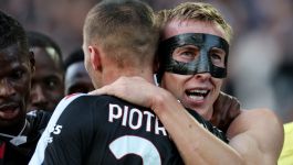 Zbliżenie na dwóch piłkarzy w czarno-białych strojach z numerami na plecach, celebrujących gola uściskiem. Piłkarz na pierwszym planie, odwrócony plecami, ma na koszulce widoczny napis "PIOTR" i dużą cyfrę "2". Piłkarz stojący po prawej stronie (prawdopodobnie Adam Buksa, ze względu na informacje z wyszukiwarki o jego kontuzji i masce) jest zwrócony w stronę aparatu, ma na twarzy czarną maskę ochronną i szeroko się uśmiecha z otwartymi ustami, patrząc lekko w górę i w prawo.