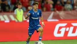 Pablo Rodriguez, hiszpański piłkarz Lecha Poznań, w niebieskiej koszulce klubowej z wyraźnym logo "SUPERBET" na środku, biegnie z piłką na boisku podczas meczu. Ma kręcone, brązowe włosy, intensywne spojrzenie i opaskę na lewym nadgarstku. Na pierwszym planie widać jego muskularne nogi i białe buty sportowe. W tle, za czerwoną bandą reklamową z logo "G-D", widać niewyraźne sylwetki kibiców na trybunach.