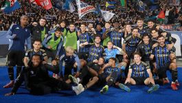 Na zdjęciu widzimy eksplozję radości – piłkarze i sztab drużyny Pisa SC świętują historyczne zwycięstwo na tle niebieskiego boiska, otoczeni falą entuzjastycznych kibiców. Zawodnicy w charakterystycznych niebiesko-czarnych pasiastych koszulkach z napisem „Central” na piersi tworzą zwartą grupę – część z nich klęczy lub siedzi z przodu, inni stoją z uniesionymi rękami, uśmiechnięci, objęci, niektórzy z wyciągniętymi flagami. W tle trybuny pulsują życiem (fot. Getty Images)
